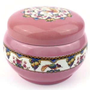 May include: A pink ceramic jar with a lid, featuring a decorative band with a bird, fruit, and floral design. The lid also has a floral and bird pattern. The jar is a soft pink color.