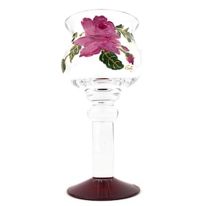 May include: A clear glass candle holder with a burgundy base and a long stem. The top of the holder is painted with a pink rose and green leaves. The initials "GA" are painted on the side of the glass.