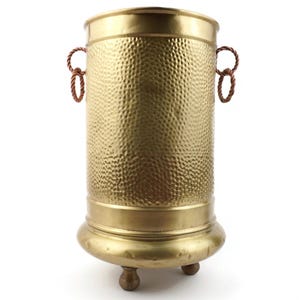 May include: A gold-colored, hammered metal umbrella stand. The cylindrical stand has a textured surface, a rounded base with three small feet, and two rope handles. The stand is designed to hold umbrellas and canes.