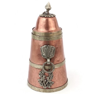 May include: A copper-colored metal container with a lid and decorative silver-colored accents. The container has a cylindrical shape with a flared base. It features a floral emblem and a decorative ring with birds.