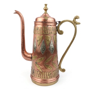 May include: A copper-colored coffee pot with a long, curved spout and a decorative handle. The pot features brass and silver-colored geometric and leaf-like patterns. The lid is topped with a gold-colored finial. The pot is likely vintage or antique.