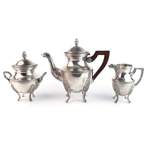 May include: A three-piece silver tea set comprising a teapot, a lidded sugar bowl, and a creamer. Each piece features ornate leaf-shaped feet, decorative bands, and a dark brown wooden handle on the teapot. The teapot has a bird-shaped spout.