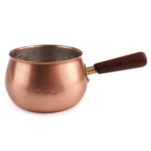 Pode incluir: Uma panela redonda de cobre com um exterior metálico polido e um revestimento interior prateado. A panela possui um único cabo horizontal feito de madeira castanha escura, fixado na lateral do recipiente com um encaixe metálico cor de latão.