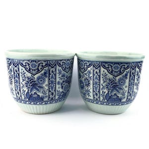 Könnte beinhalten: Zwei passende Keramik-Blumentöpfe mit blau-weißem Blumenmuster. Die Töpfe haben eine runde Form mit geripptem Boden und dekorativem Rand. Das Blumenmuster umfasst verschiedene Blumen und Blätter.