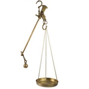 May include: A brass hanging balance scale featuring a long graduated beam with a sliding counterweight, a central suspension hook, and a single circular weighing pan suspended by three thin metal chains attached to a small hook at the top.