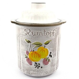 May include: A white ceramic Rumtopf pot with a lid. The pot features a painted design of yellow and orange fruit with green leaves and the word "Rumtopf" in script. The rim and lid are brown.