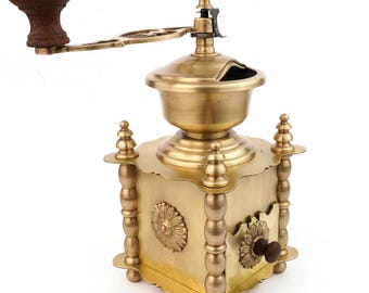 May include: A vintage brass coffee grinder with a wooden handle and knob. The grinder features ornate floral designs and decorative posts. The top has a small opening for adding coffee beans.