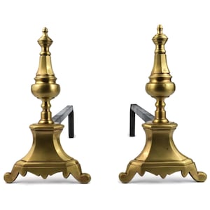 May include: A pair of antique brass and metal fireplace andirons. The andirons have a decorative, ornate design with a gold finish. The base is a detailed, curved shape, and the upright portion has a bulbous design. The metal support is black.