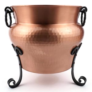 May include: A copper cauldron with a hammered finish, featuring a wide rim and two twisted rope handles. The pot is supported by a black wrought iron stand with decorative, curved legs. The pot's diameter is approximately 30 cm.