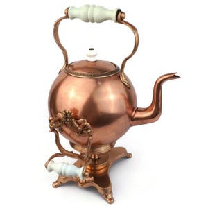 May include: A copper-coloured, spherical tea kettle with a curved spout and a matching stand. The kettle has a white handle and a white knob on the lid. The stand features a decorative floral design.