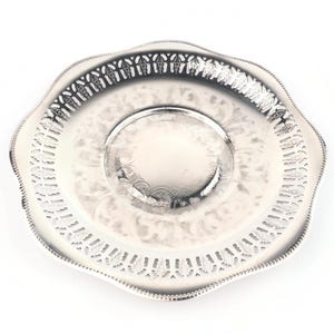 Puede incluir: Un plato decorativo plateado con borde festoneado e intrincados patrones florales y geométricos. El plato tiene un centro elevado y un borde calado, creando un diseño detallado y elegante.
