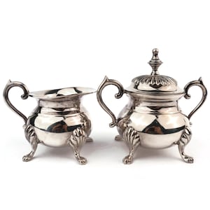 May include: A silver-colored creamer and covered sugar bowl set. The creamer has a curved handle and a scalloped rim. The sugar bowl has a lid with a decorative finial and matching handles. Both pieces have ornate, claw-footed bases.