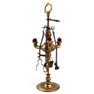 May include: A vintage brass oil lamp with a circular base and a tall, ornate design. The lamp features three spouts with red accents, hanging chains, and small tools. The top has a handle and a small tray.