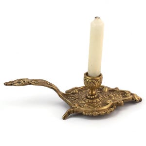 Puede incluir: Candelabro de latón ornamentado con una vela blanca encendida. El candelabro presenta un diseño floral detallado y un mango largo y curvo. La vela es de tamaño estándar y la pieza tiene una estética vintage.