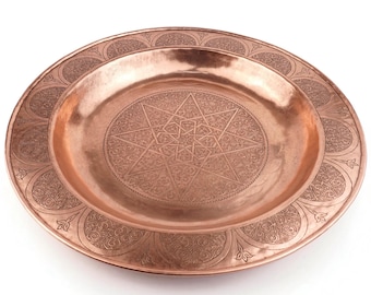 May include: A copper-colored decorative plate with an intricate star and floral pattern. The plate has a raised center and a wide, detailed border. The design is etched into the metal, creating a textured surface. The plate is likely used for display or serving.