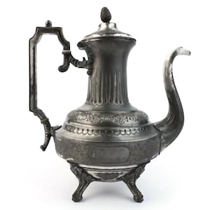 May include: Antique silver-colored coffee pot with a decorative handle and spout. The pot has a fluted body, ornate floral detailing, and rests on three decorative feet. The lid is topped with a small, cone-shaped finial. A vintage kitchenware item.