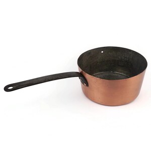 Op de afbeelding: Een koperen steelpan met een lange, zwarte handgreep. De pan heeft een afgeronde vorm en een klein gaatje bij de rand. De handgreep is aan de zijkant geklonken.