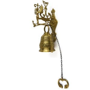 Puede incluir: Una campana de latón montada en la pared con un diseño ornamentado. La campana presenta una figura de ángel, adornos decorativos y una cadena con un tirador en forma de corazón. La campana está grabada con la palabra "TANGIT" y figuras de animales.