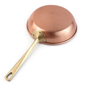 Pode incluir: Uma frigideira redonda de cobre com acabamento liso e polido e um cabo longo e fino de latão. O cabo está fixado na lateral da frigideira com uma placa de metal curva com três rebites visíveis e inclui um orifício na extremidade.