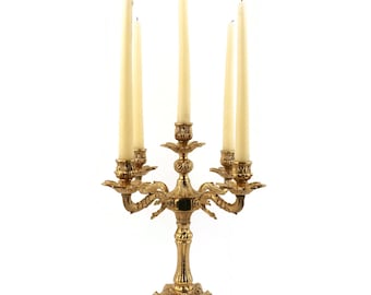 May include: A gold-toned metal candelabra with five candle holders and ornate details. The candelabra has a base with a decorative design and a central stem with five arms that curve outwards. Each arm has a candle holder with a decorative design. The candelabra is a decorative piece that can be used to hold candles.