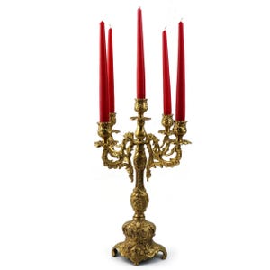 May include: A gold-colored candelabra with five red taper candles. The candelabra has an ornate, detailed design with a round base and multiple arms to hold the candles. The candles are a vibrant red color.