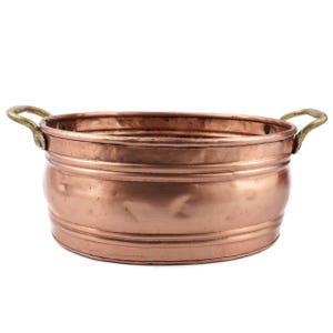 Puede incluir: Un cuenco redondo de color cobre con dos asas de latón. El cuenco tiene una parte superior ancha y abierta y un cuerpo ligeramente curvado. El exterior presenta bandas horizontales. Las asas están fijadas a los lados. El diámetro es de unos 25 cm.
