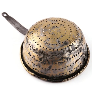 May include: A round, dome-shaped brass colander featuring a pattern of small, evenly spaced drainage holes across its surface. The metal shows signs of wear with dark, tarnished patches and a long, narrow, dark-colored metal handle attached.