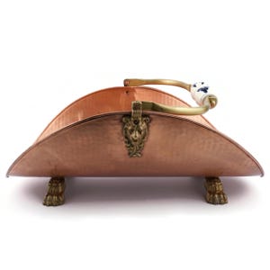 Puede incluir: Un leñero de cobre con asa de latón y detalle de cabeza de león. El asa tiene un agarre de cerámica blanca con detalles florales azules. El leñero descansa sobre patas de garra de latón, mostrando un diseño antiguo.