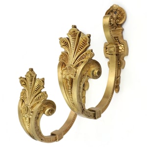 Puede incluir: Dos ganchos de pared dorados y ornamentados con un diseño clásico y vintage. Cada gancho presenta una parte superior decorativa en forma de abanico y un gancho curvado. Los ganchos son de metal y están montados sobre un fondo blanco.
