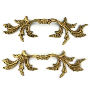 Op de afbeelding: Twee sierlijke, goudkleurige ladehandgrepen met een bloemen- en bladerontwerp. De handgrepen hebben een gebogen vorm met gedetailleerde versieringen, perfect om een vintage touch aan meubels toe te voegen. De hardware is van metaal.
