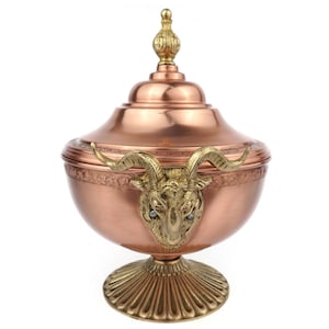 May include: A copper-colored decorative container with a lid, featuring a gold-colored ram's head with blue accents. The container has a pedestal base and a decorative finial on top. The design is ornate and detailed.