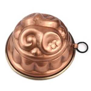 Puede incluir: Un molde decorativo de color cobre con una parte superior redondeada y una base circular. El molde presenta patrones ornamentados y arremolinados y un pequeño asa negra. La base tiene un borde color latón.