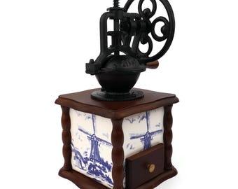 May include: A vintage-style coffee grinder with a black cast iron mechanism atop a wooden base. The base features blue and white windmill tile panels and a small drawer. The grinder has a decorative handle and a detailed design.