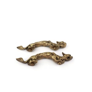 Op de afbeelding: Twee antieke messingkleurige kastgrepen in de vorm van gestileerde fabeldieren. Elke greep heeft een gedetailleerd ontwerp met een gebogen lichaam, een kop met hoorns en een staart. De grepen zijn gemonteerd op een witte achtergrond.