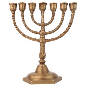May include: A gold-colored menorah with seven candle holders. The base is square and tiered, with a decorative floral design on the central stem. The arms curve upwards, each holding a cup-shaped candle holder.