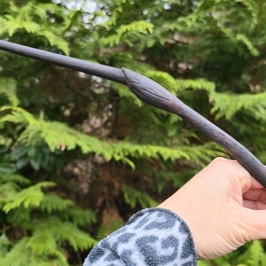 May include: A dark brown wooden wand with a textured handle and a carved design near the top. The wand is held in a hand against a backdrop of green foliage. The wand appears to be approximately 14 inches long.