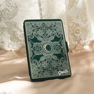 以下が含まれることがあります： 濃い緑色の携帯電話ケース。金色の花と月のデザインが施されています。「Giselle」という名前と三日月が特徴です。背景には柔らかいニュートラルカラーの生地が使われています。