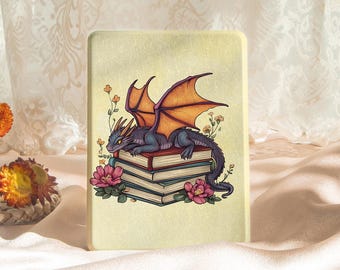 May include: A rectangular, cream-colored decorative item featuring a cartoon dragon with purple scales and orange wings, perched atop a stack of books. Pink flowers and small orange blossoms surround the dragon and books.