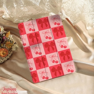 May include: A red and pink checkered tablet case featuring bows and cherries. The case has a repeating pattern of red bows and cherries on a pink and red checkered background. The case is on a satin surface.