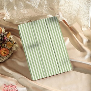 Puede incluir: Una funda para tableta con un patrón de rayas verdes y blancas. La funda está abierta, revelando el interior. El fondo presenta una cesta de flores secas y tela drapeada.