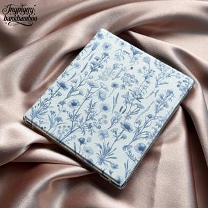 May include: A tablet case with a white background and a blue floral pattern. The case is rectangular and has rounded corners. The case is lying on a pink satin surface. The text "Ingpiggy bankbamboo" is in the upper left corner.