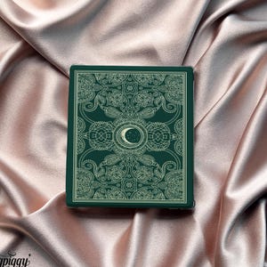 May include: A dark green rectangular object featuring a detailed, symmetrical pattern and a crescent moon design. It rests on a backdrop of pink satin fabric.