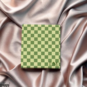 Könnte beinhalten: Ein quadratischer Gegenstand mit einem grün-cremefarbenen Schachbrettmuster und den Initialen "LM" unten rechts. Er liegt auf einem rosafarbenen Satinstoff. Das Logo "Ingpiggy bankbamboo" ist unten links zu sehen.