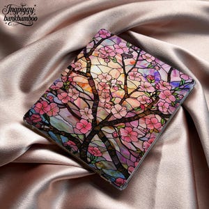 May include: A clear case featuring a stained-glass design of a flowering tree with pink blossoms. The branches are dark brown, and the background has shades of orange, yellow, and purple. The case is on a satin surface.