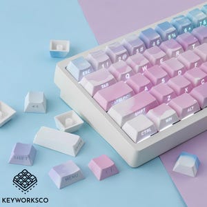 May include: A white mechanical keyboard with pastel-colored keycaps. The keycaps transition from blue to pink, with white lettering. Several loose keycaps are scattered around the keyboard. The brand logo "KEYWORKSCO" is visible in the bottom left corner.