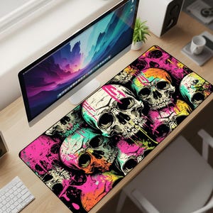 May include: A black desk mat with a striking design of skulls in white, orange, pink, yellow, and teal. The skulls are overlaid with paint splatters, creating a bold and colourful aesthetic. This desk mat is designed for computer use.