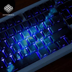 May include: A close-up of a mechanical keyboard with custom keycaps. The keycaps feature a galaxy design with blue and white colors. The keyboard has blue LED backlighting. The brand name "KEYWORKSCO." is visible in the upper left corner.