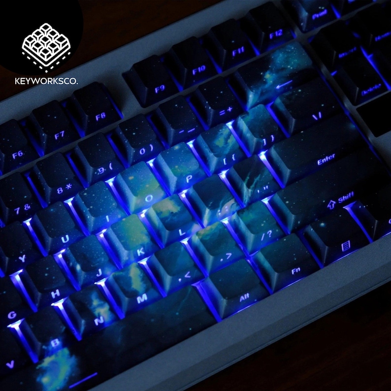May include: A close-up of a mechanical keyboard with custom keycaps. The keycaps feature a galaxy design with blue and white colors. The keyboard has blue LED backlighting. The brand name "KEYWORKSCO." is visible in the upper left corner.