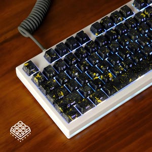 May include: A white mechanical keyboard with black keycaps speckled with yellow. The keyboard has a grey coiled cable and is lit with blue underglow. The keycaps are arranged in a standard QWERTY layout.