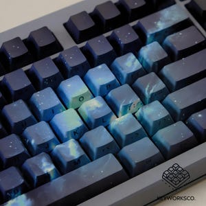 May include: A close-up of a mechanical keyboard with custom keycaps. The keycaps feature a blue and white galaxy design, with a mix of dark blue and lighter blue hues. The keyboard has a dark gray frame and the brand logo is visible.